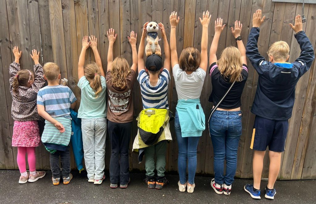 Acht Kinder stehen mit dem Rücken zur Kamera vor einer Bretterwand. Alle halten ihre Hände in Jubelpose in die Höhe.